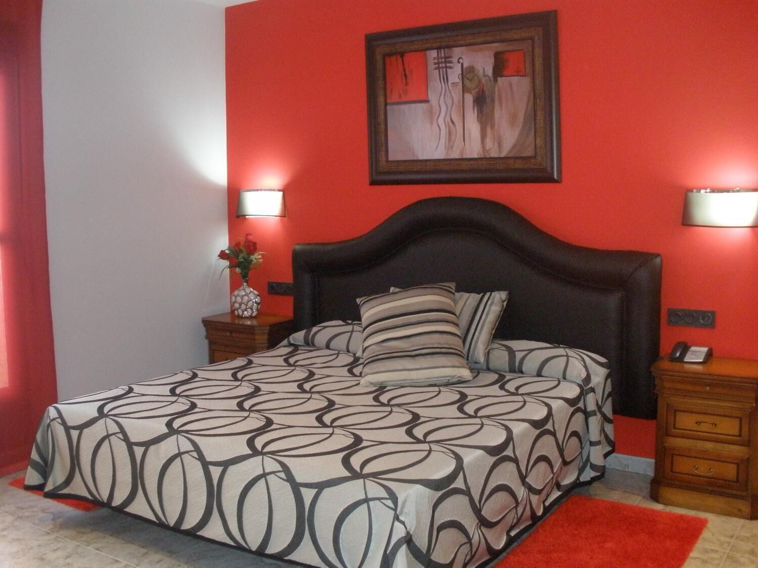 A bedroom with a bed, two bedside tables, a nightstand, a red wall, and a painting.