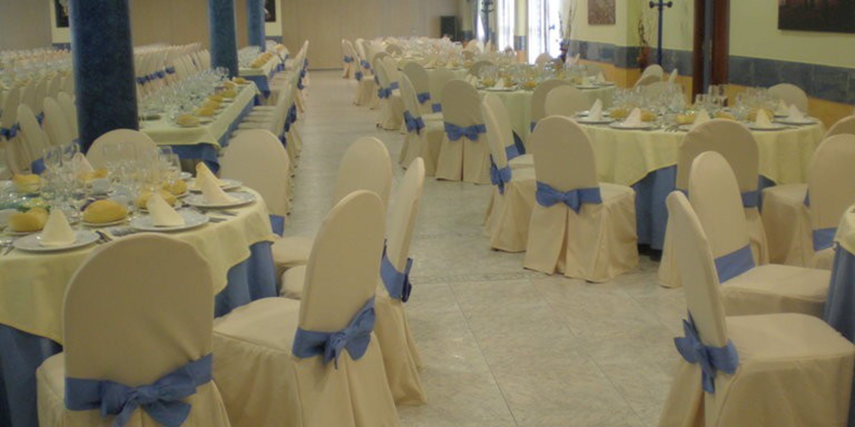 Uma sala de banquetes com fileiras de mesas montadas para um evento formal, cada uma com toalhas brancas e fitas azuis nas cadeiras.