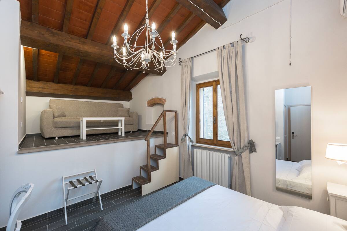 Ein Schlafzimmer mit Bett, Nachttisch, Kommode, einem Fenster mit Vorhängen, einer Chandelierleuchtre und einer Treppe. In einem angrenzenden Raum befindet sich eine Couch.