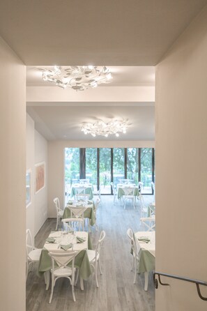 A dining area with white tables and chairs, a chandelier, and large windows allowing natural light. - Hotel le Macine (Montaione)
