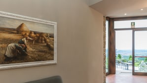 A hallway with a large painting of a rural scene, a cushioned bench, and a view of the outdoors through a glass door. - Hotel le Macine (Montaione)