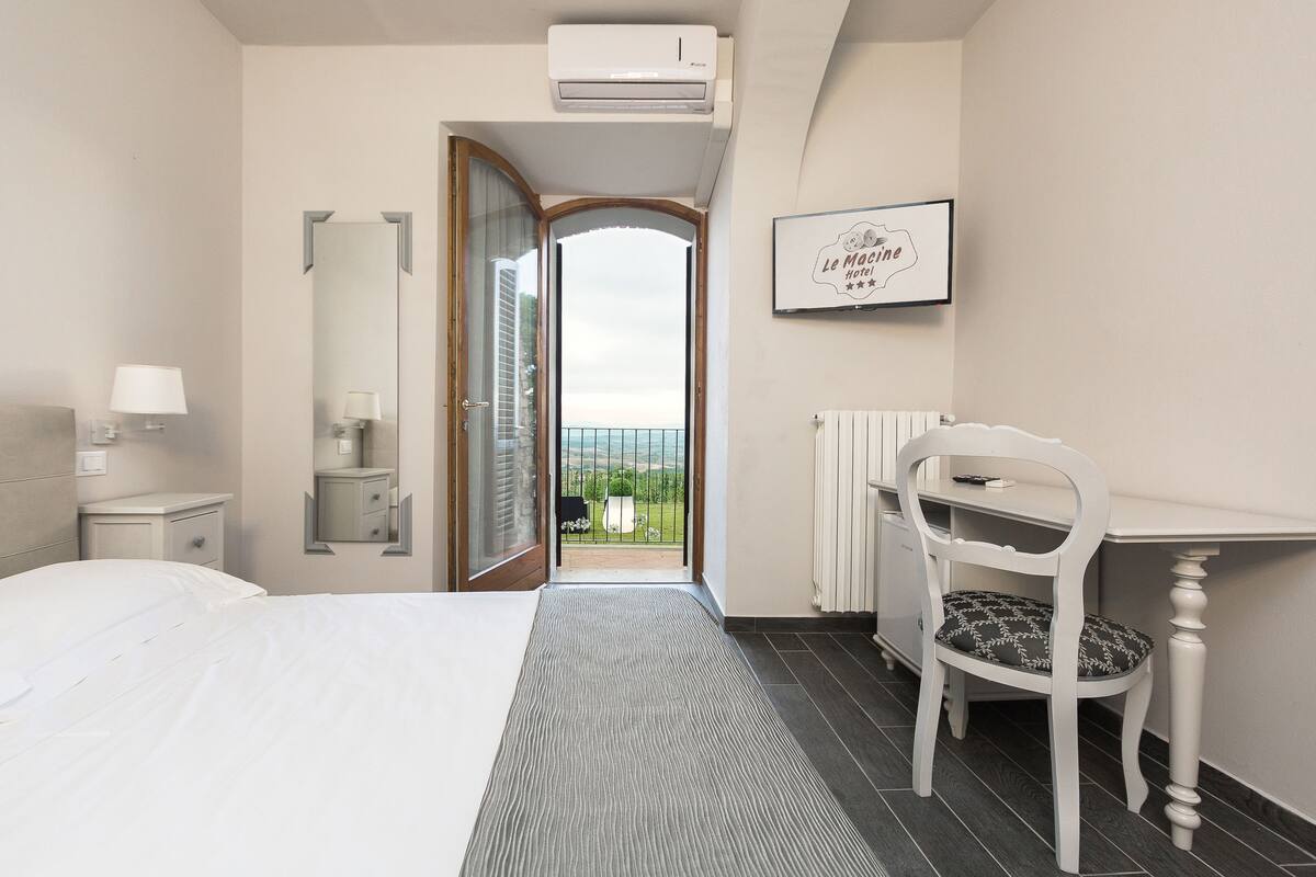Ein Hotelzimmer mit einem Bett, einem Schreibtisch, einem Sessel und einem Spiegel, mit Blick ins Freie.