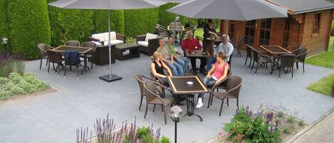 Un patio al aire libre con mesas y sillas bajo sombrillas, rodeado de setos cuidados y diversas plantas.