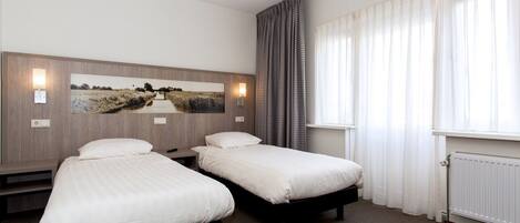 A hotel room with two single beds, a large landscape painting on the wall, a window with sheer curtains, and a wall-mounted light fixture.