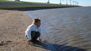 A child crouching by a riverbank with wind turbines in the background. - Hotel Ekamper (Roodeschool)