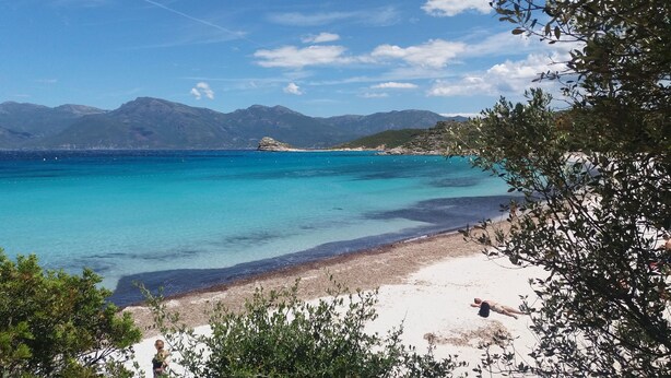 Une plage à l’eau turquoise et limpide, un rivage sablonneux et des montagnes qui se dessinent à l’horizon.