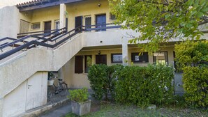 Un bâtiment à deux étages avec un escalier menant à un balcon. On peut voir des plantes en pot et un vélo près de l’entrée.