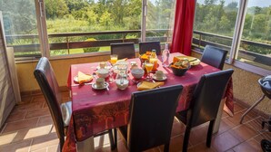 Une table à manger dressée avec des aliments pour le petit-déjeuner, des chaises, et une vue sur la verdure grâce à de grandes fenêtres.