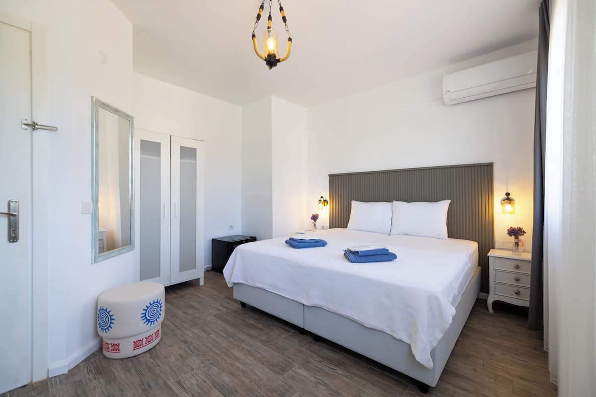 A modern hotel room with a large bed, bedside lamps, a mirror, a small stool, and an air conditioning unit.