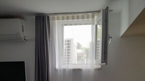A room with a window, a flat-screen TV mounted on the wall, a power outlet, and a small table with a chair. - Elvin Bozcaada (Bozcaada)