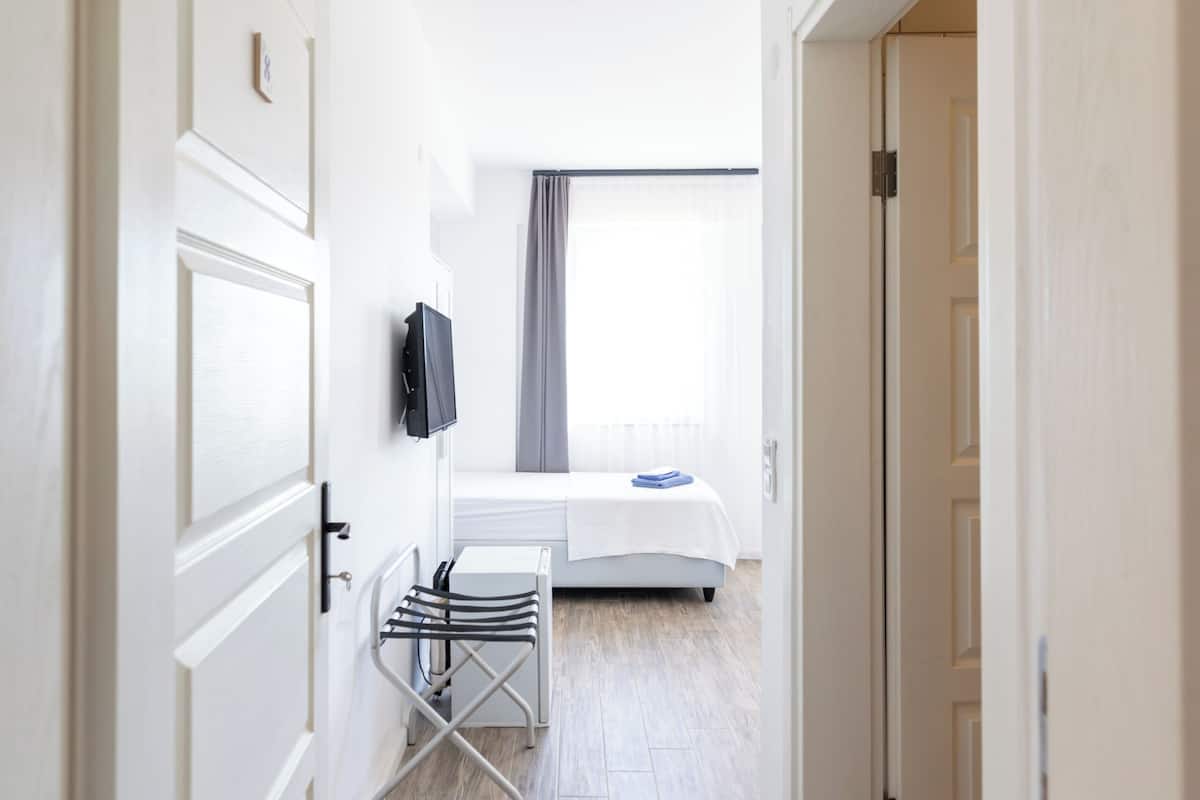 A hotel room with a bed, a TV mounted on the wall, a window with curtains, and a wooden floor.