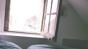 Ein Zimmer mit einem Einzelbett, einem Fenster mit Blick auf Gebäude und einem Heizkörper an der Wand.