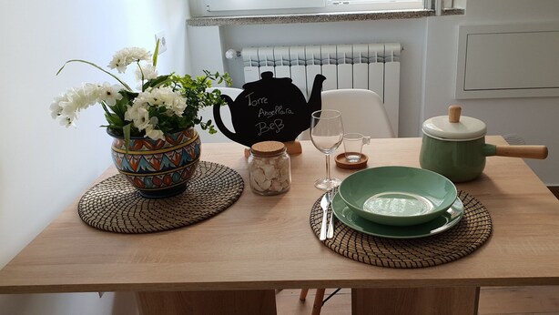 Una mesa de madera con un jarrón de flores blancas, una tetera, un tazón verde y un tarro de conchas marinas.