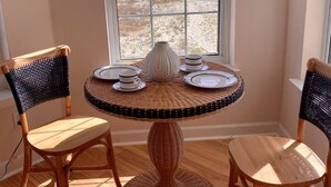 Une pièce d’angle avec une grande fenêtre, une table ronde en osier sur laquelle sont disposées des assiettes et des tasses, deux chaises en osier et un plancher en bois.