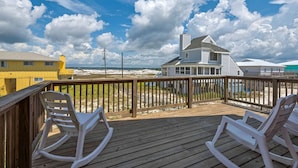 Une terrasse en bois avec deux fauteuils à bascule, une rambarde en bois et une vue sur une plage et une maison.