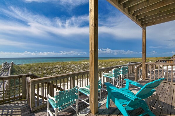 A wooden deck with turquoise chairs and a table overlooking a beach.
