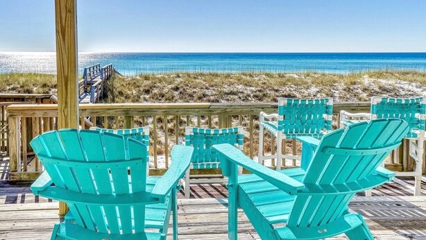 A wooden deck with turquoise Adirondack chairs and a view of the beach.