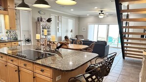 A spacious kitchen with granite countertops, wooden cabinets, and a central island. There is a dining area with a table and chairs, and a living room with a sofa and a ceiling fan.