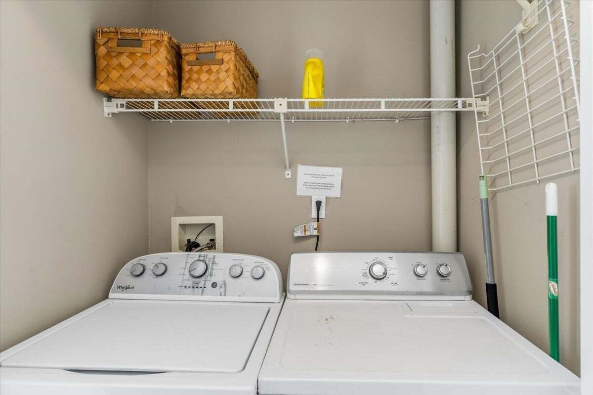 A laundry room with two white Whirlpool washers, a metal shelf with baskets, and a mop.