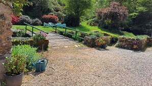 A gravel pathway leading to a wooden bridge over a stream, surrounded by lush greenery and stone walls.