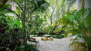 A paved garden area with a bench and a table, surrounded by lush greenery and plants.