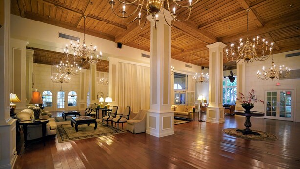 A spacious lobby with high ceilings, wooden floors, and multiple chandeliers. There are several seating areas with sofas and armchairs, and a large mirror reflecting the space.