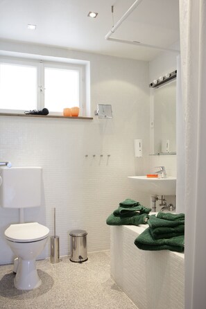 A modern bathroom with a toilet, sink, and mirror. There are green towels on the sink, a trash can, and a window above the toilet.
