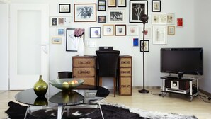 A living room with a round glass table, a black chair, a television on a stand, and a wall decorated with framed pictures. - House on Schlossberg (Tuebingen)