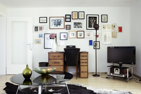 A living room with a round glass table, a black chair, a television on a stand, and a wall decorated with framed pictures.