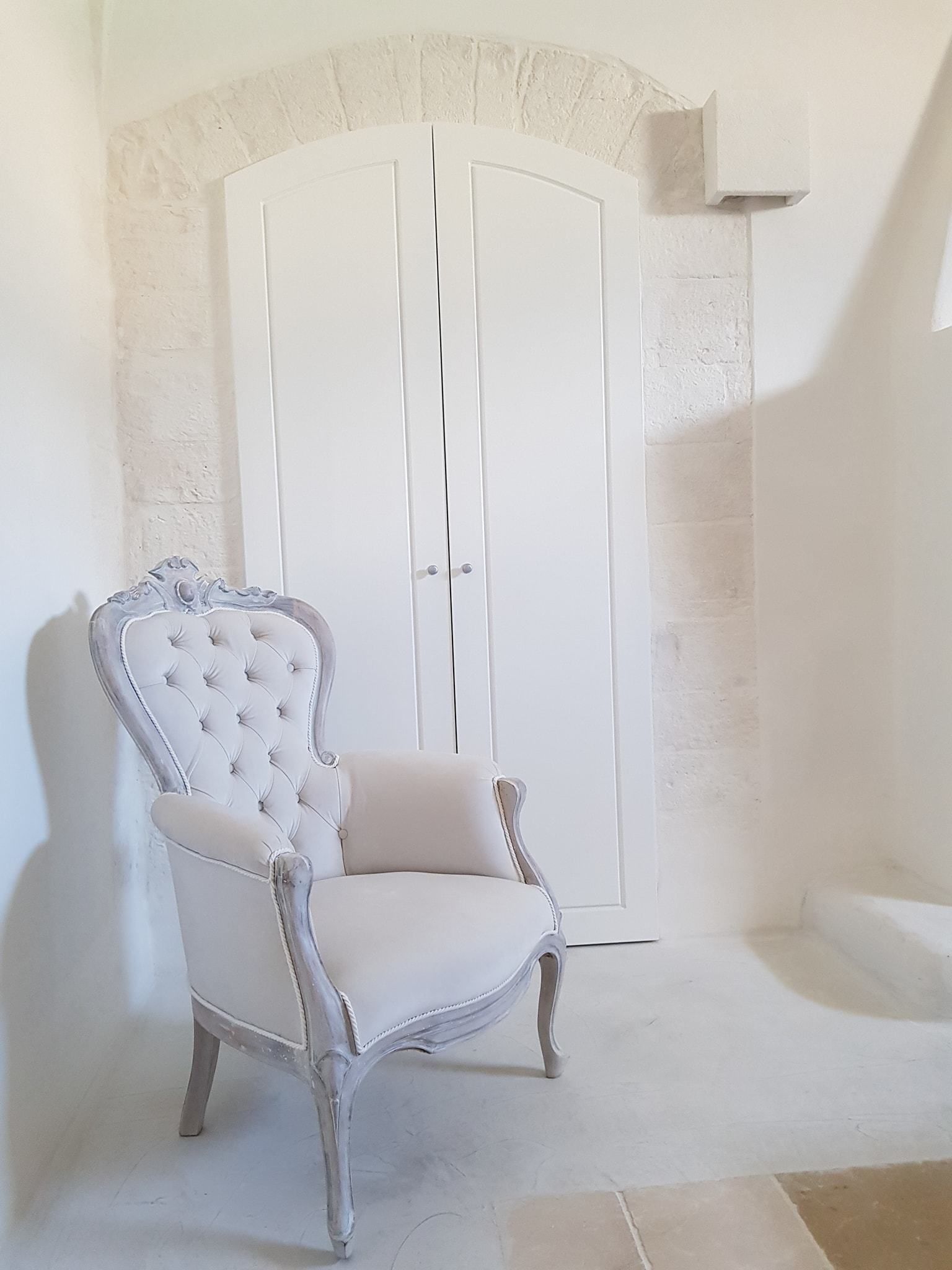Un fauteuil blanc, recouvert de tissu, doté d’un dossier capitonné et de pieds incurvés, placé devant une porte blanche surmontée d’un arc en pierre.