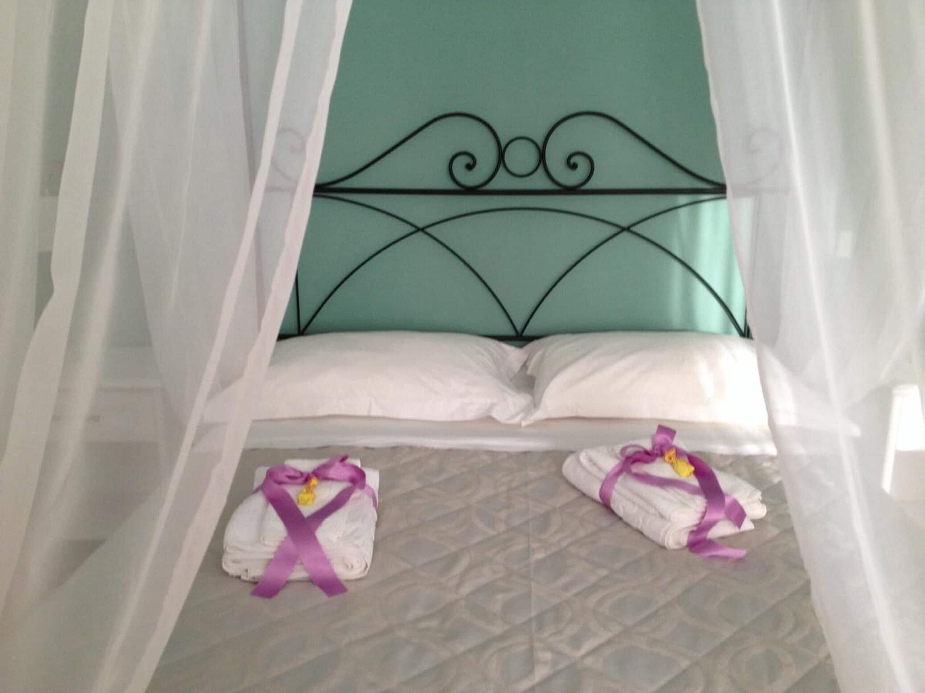 Una cama con cabecera verde azulada, almohadas blancas y dosel. Hay dos toallas con lazos morados sobre la cama.