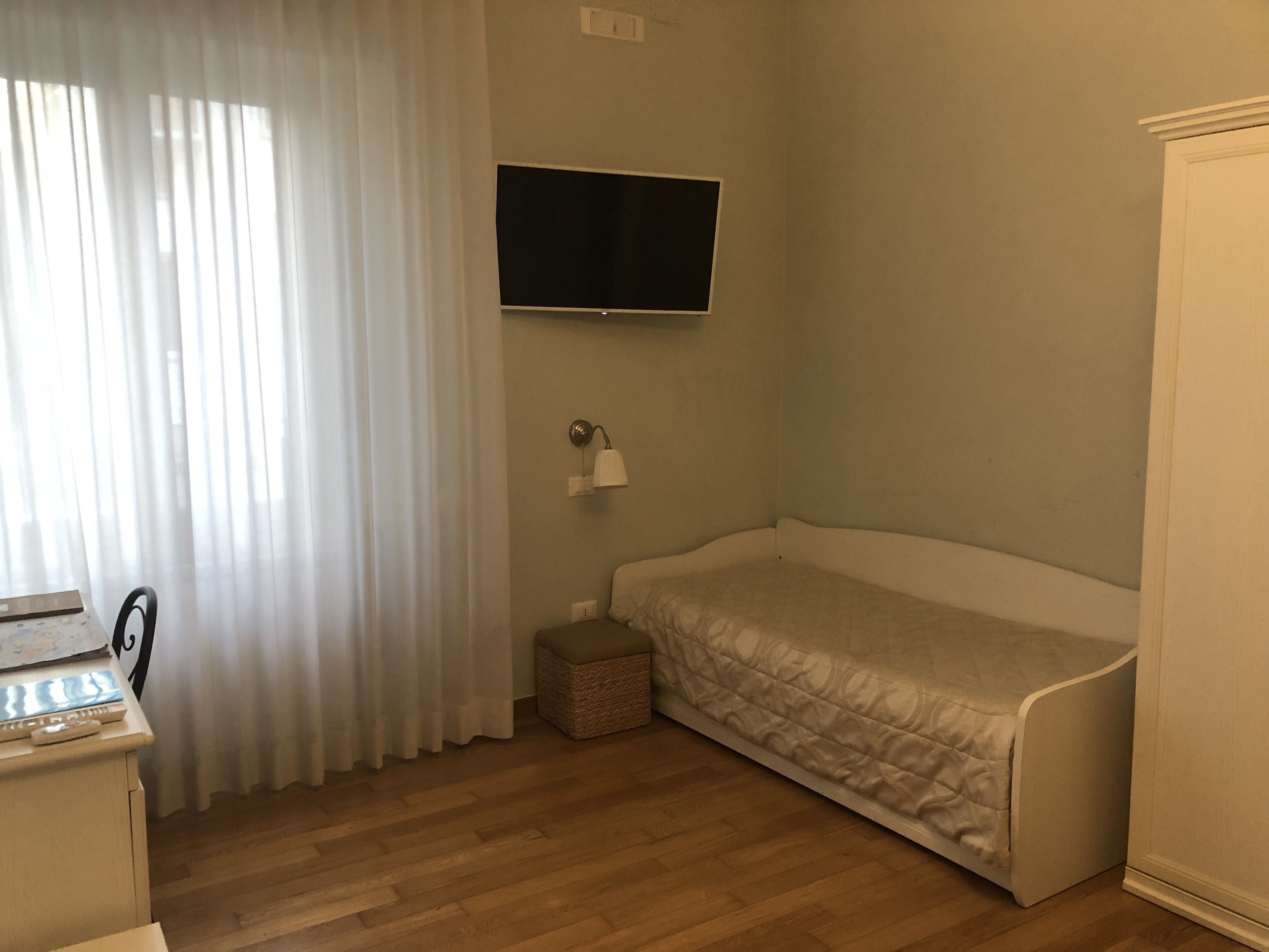 Un petit logement composé d’une seule pièce, comprenant un lit, un téléviseur fixé au mur, une table de chevet et une fenêtre avec des rideaux légers.