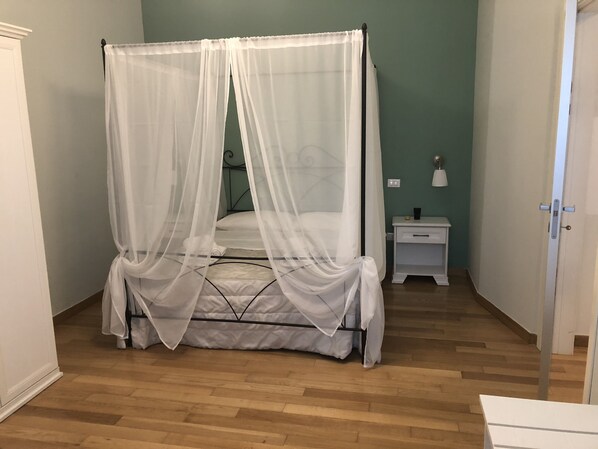 Una cama con dosel y cuatro columnas, con una colcha y almohadas blancas, una mesita de noche con lámpara, y piso de madera.