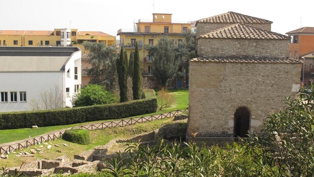 A historical site with ancient ruins, a stone building, and modern residential buildings in the background. - B&B Arco dolce Arco (Benevento)