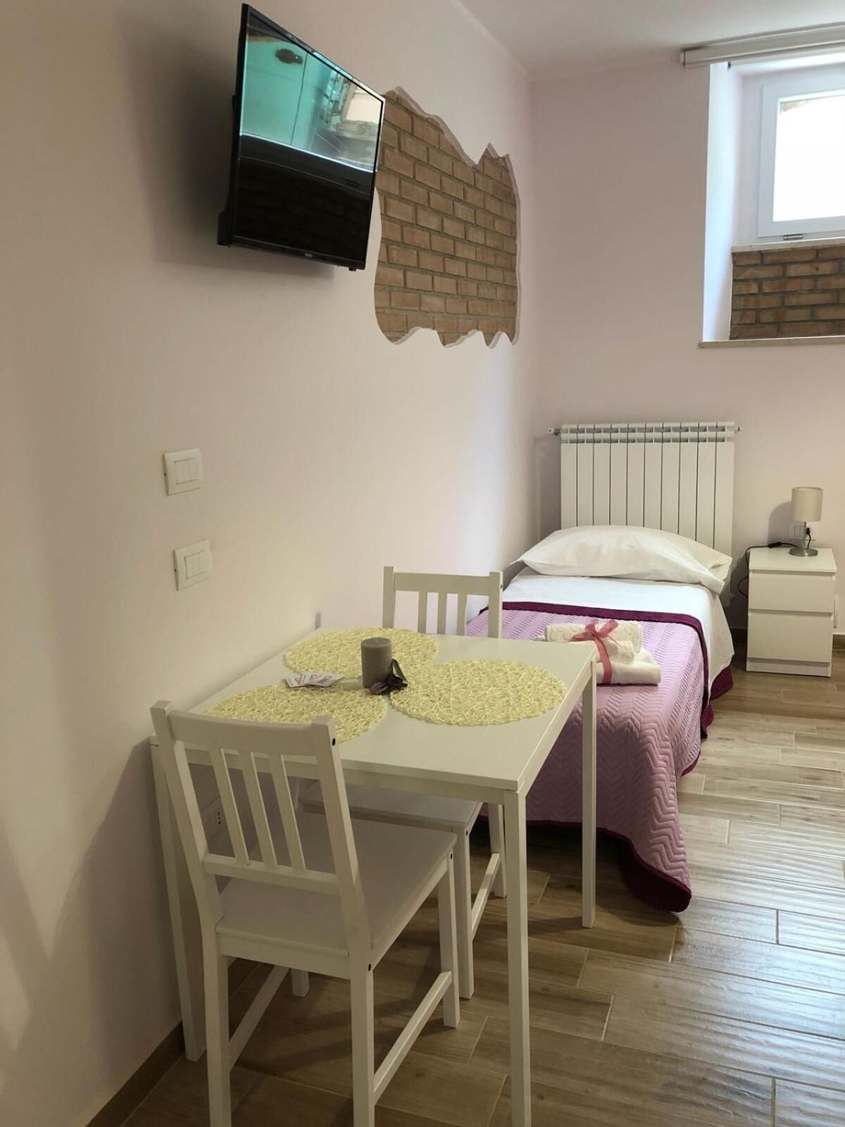 A bedroom with a bed, a bedside table, a TV mounted on the wall, and a window.