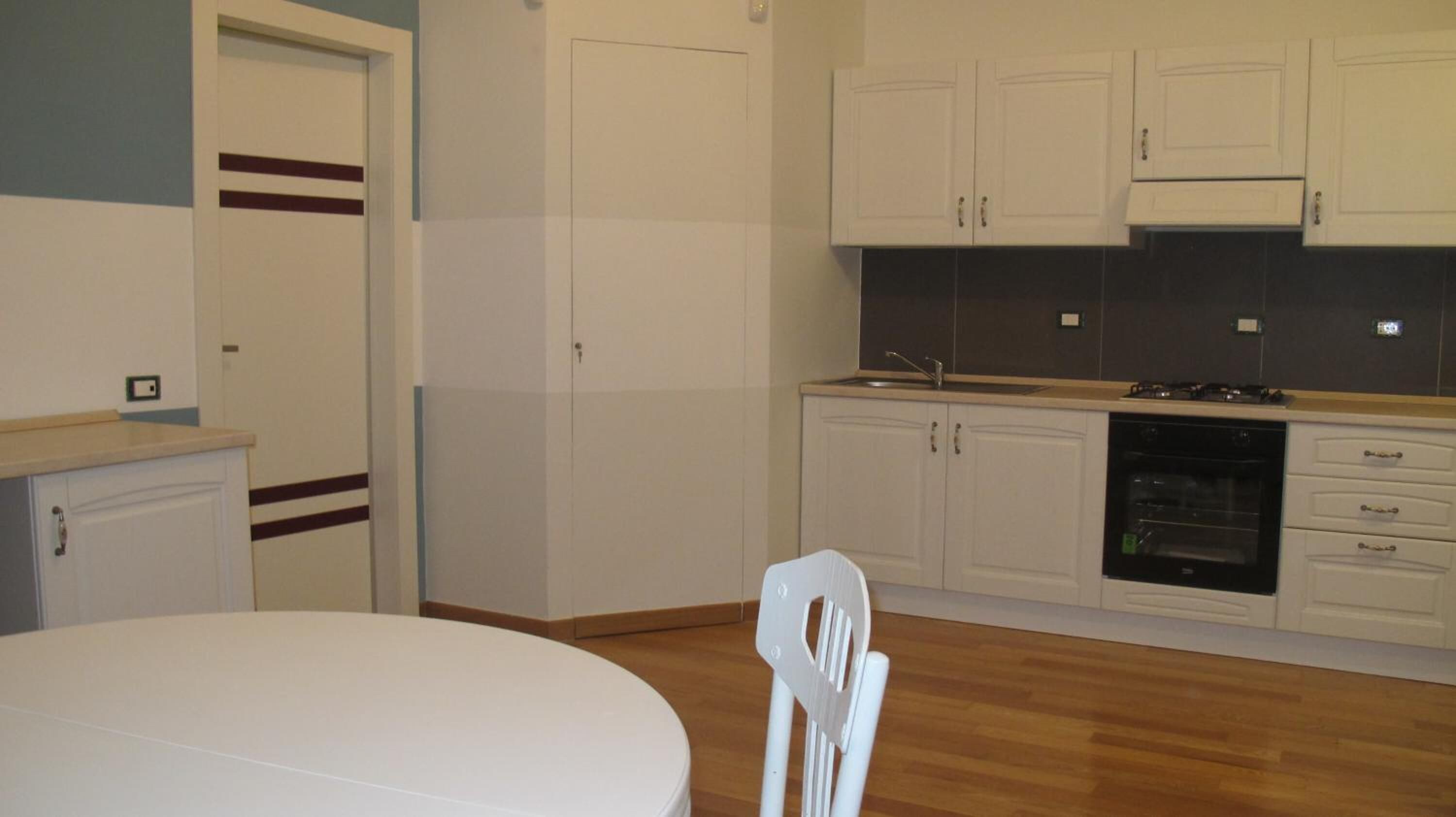 A compact kitchen with white cabinets, a stove, and a sink. There is a round table and a chair in the foreground.