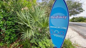 Un panneau bleu en forme de planche de surf, portant les inscriptions « Heidiway » et « 30a vacation », et entouré de verdure.