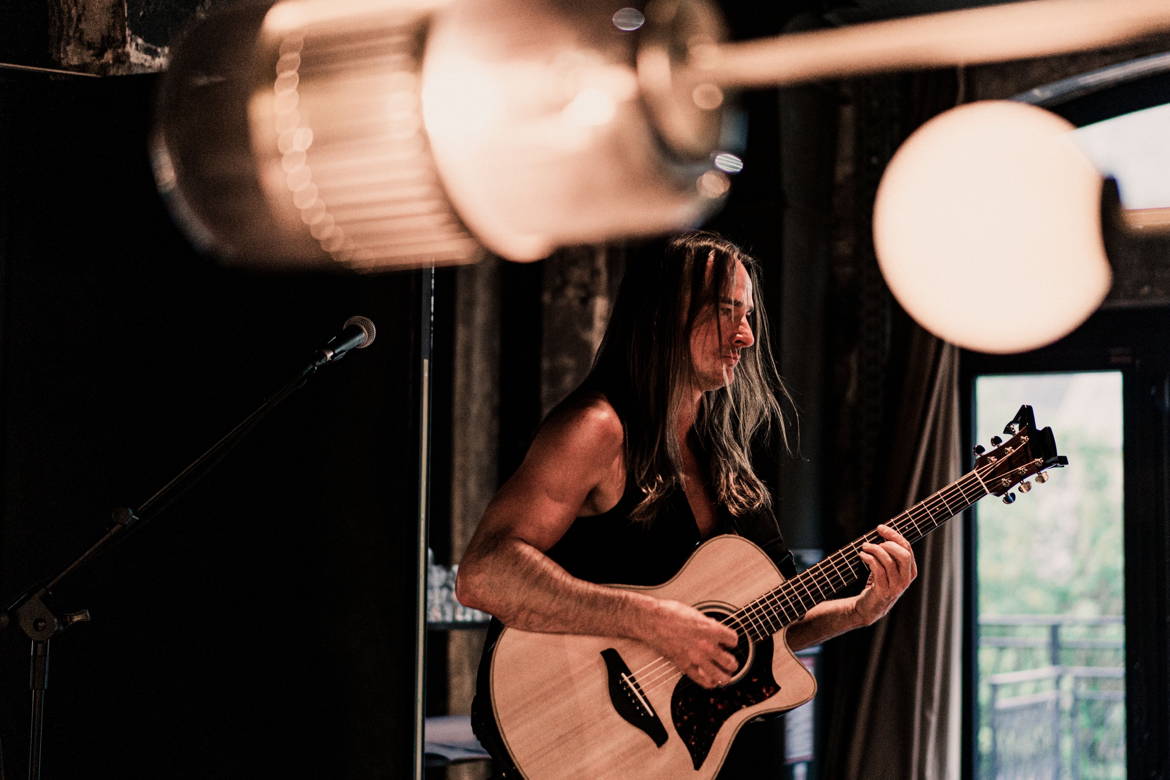 A person playing an acoustic guitar in a dimly lit room with a microphone stand and a window in the background.