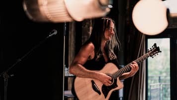 A person playing an acoustic guitar in a dimly lit room with a microphone stand and a window in the background.