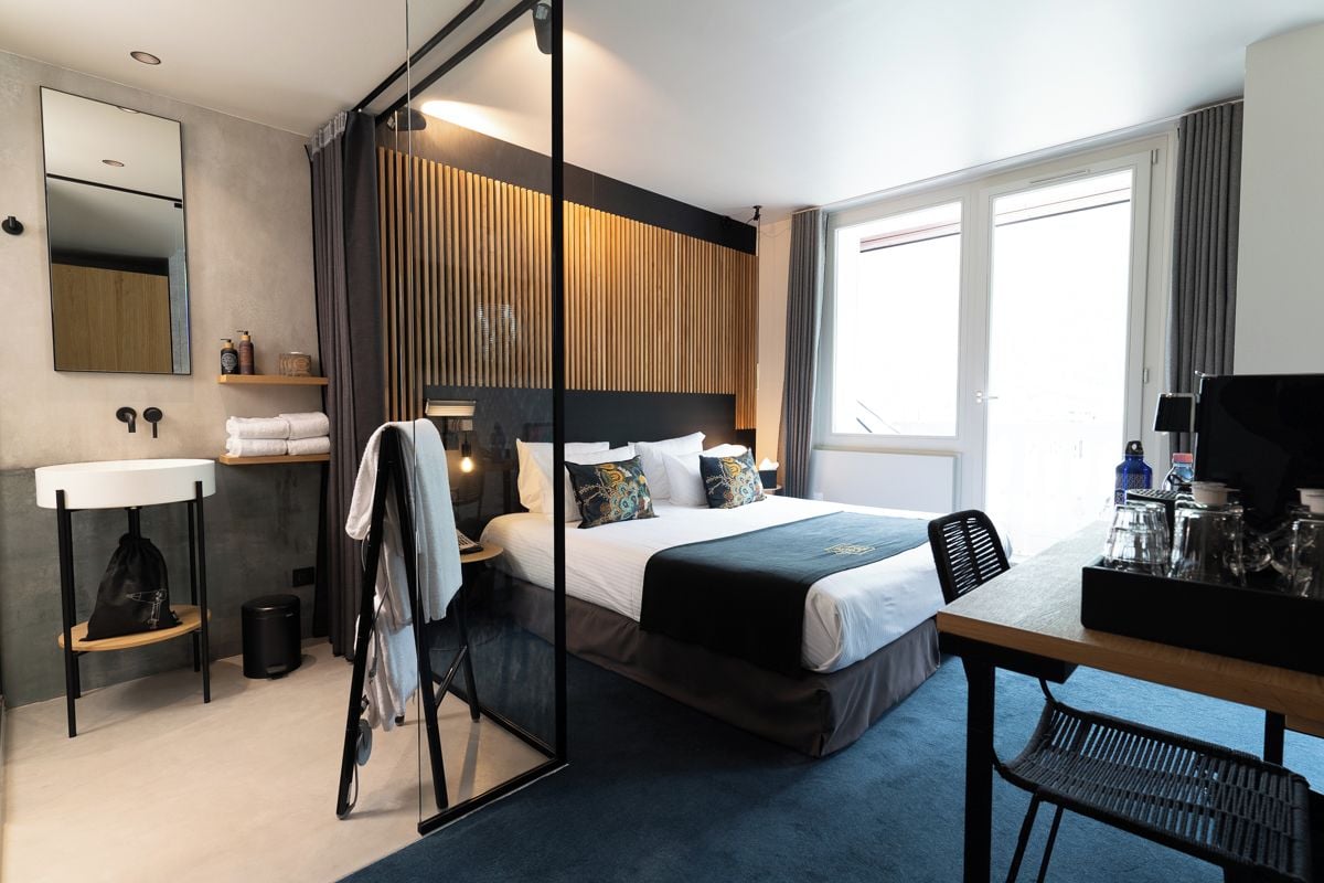 Una habitación de hotel moderna con cama, mesita de noche, espejo y vistas al exterior.