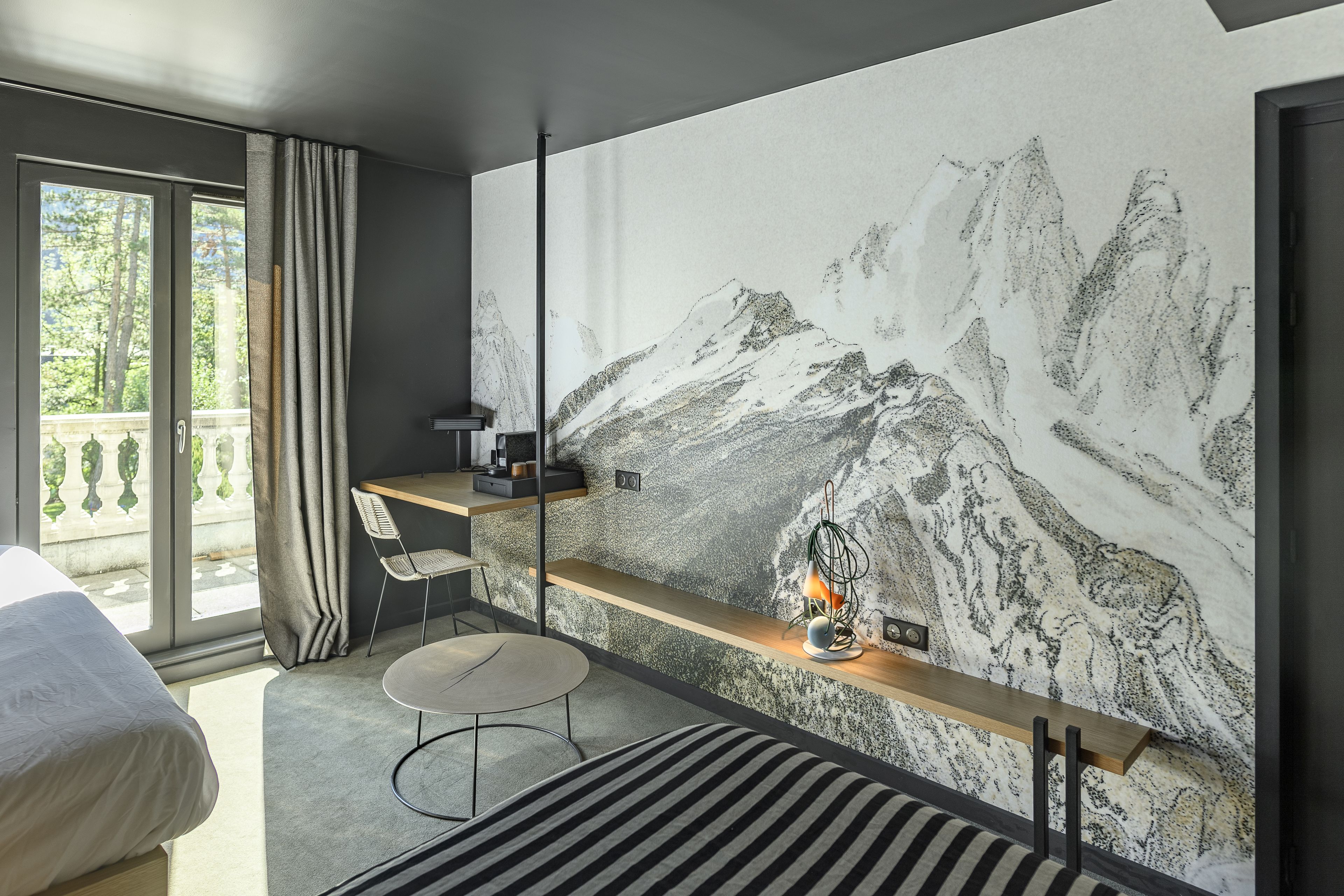 Una habitación de hotel moderna con una cama grande, un escritorio con una silla, una pequeña mesa redonda y una pared decorada con una pintura de montañas.