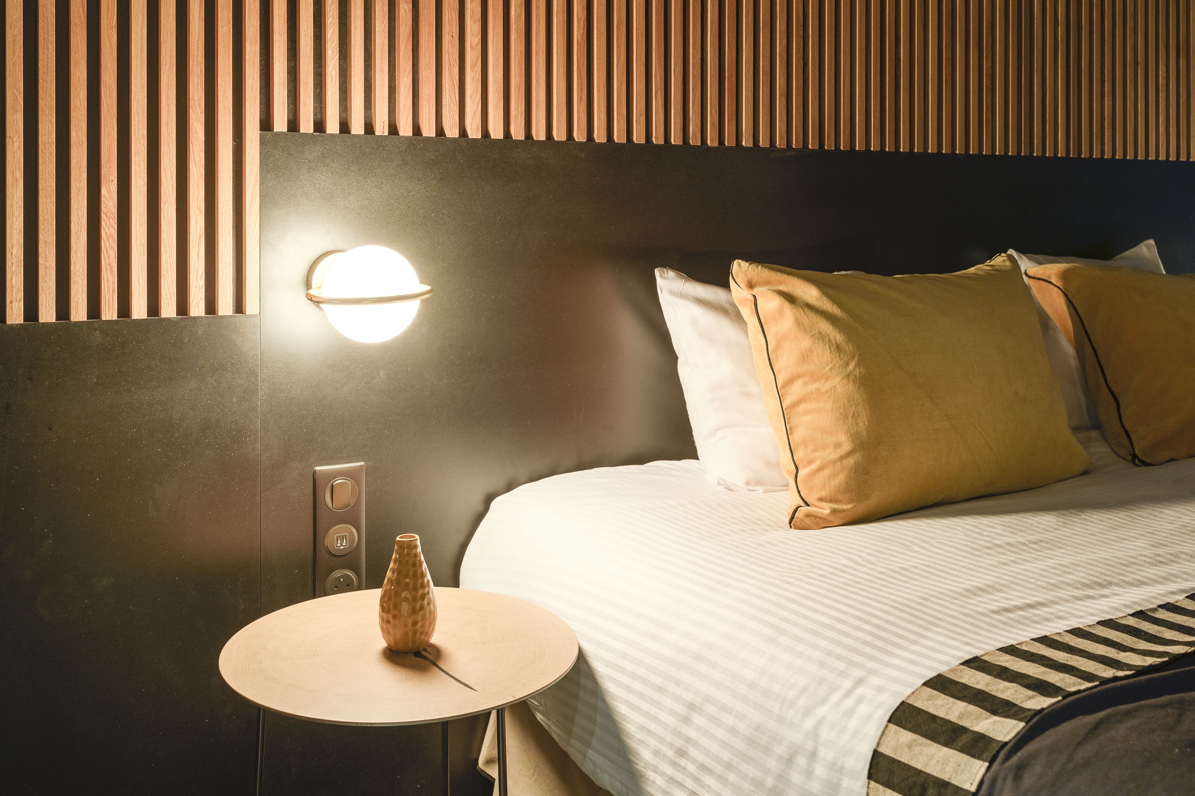 A hotel room with a bed, a bedside table with a vase, a wall-mounted light, and a wooden panel design.