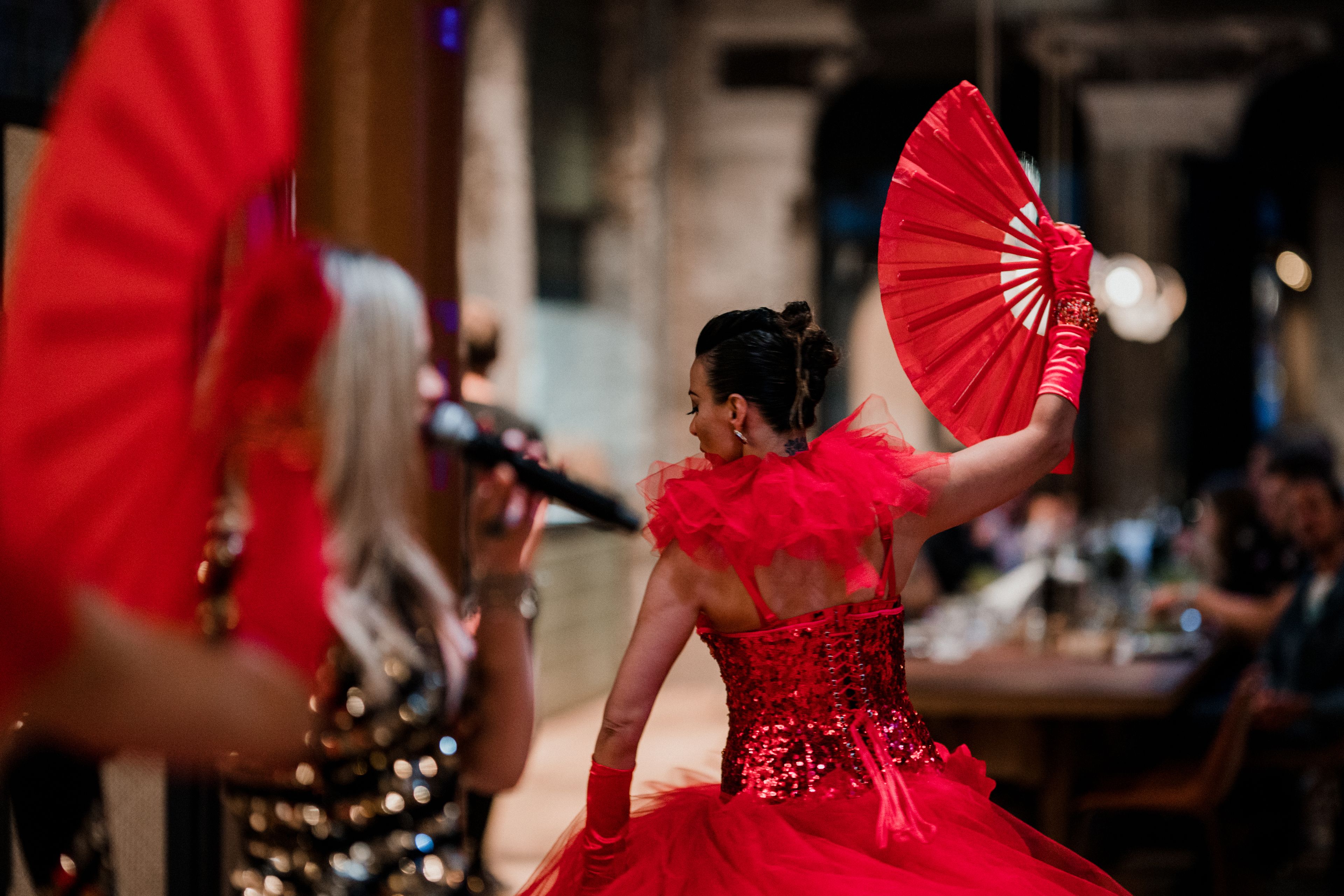 A person in a red dress holding a fan, with another person in a sequined dress in the foreground.