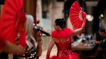 Une personne vêtue d’une robe rouge tient un éventail, et une autre personne, portant une robe à paillettes, se trouve au premier plan.