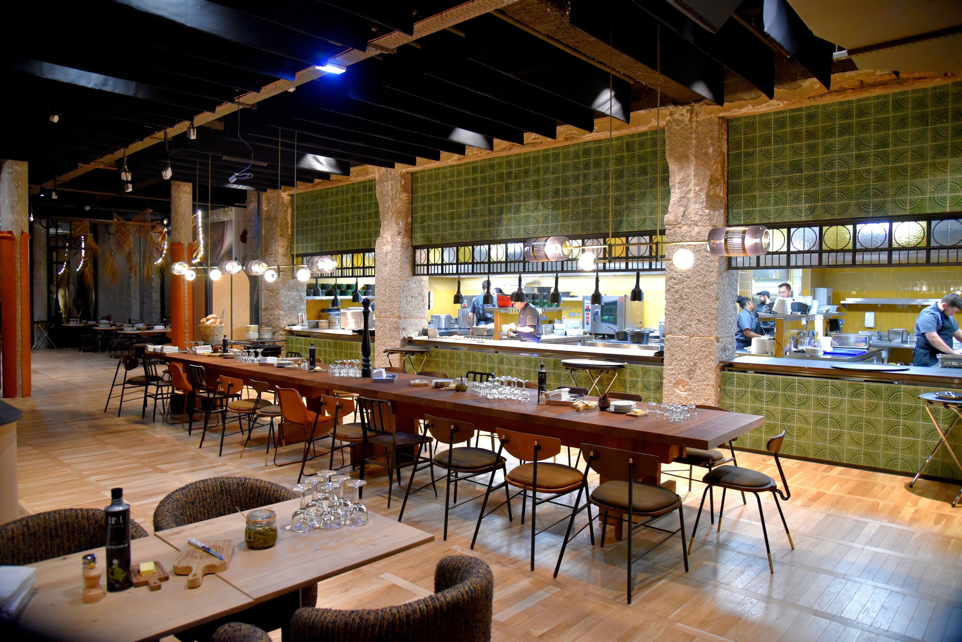 A restaurant with a long wooden table, multiple chairs, and a counter where a chef is preparing food.