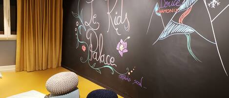 Una habitación con pared negra que presenta texto y gráficos coloridos, dibujados a mano, incluyendo una ilustración de un pájaro y las palabras "La Folie Douce Chamonix". Hay tres cojines redondos sobre un suelo amarillo.