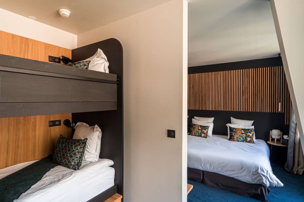Une chambre d’hôtel moderne avec un lit superposé, une tête de lit en bois, une table de chevet avec une lampe et une grande fen être avec des rideaux.