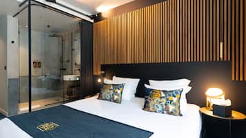 A modern hotel room with a glass shower enclosure, a wooden slatted headboard, and a neatly made bed with patterned pillows.