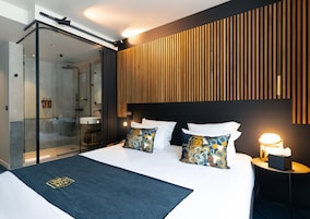 Una habitación de hotel moderna con cabina de ducha de vidrio, cabecero de madera con listones y una cama bien hecha con almohadas estampadas.