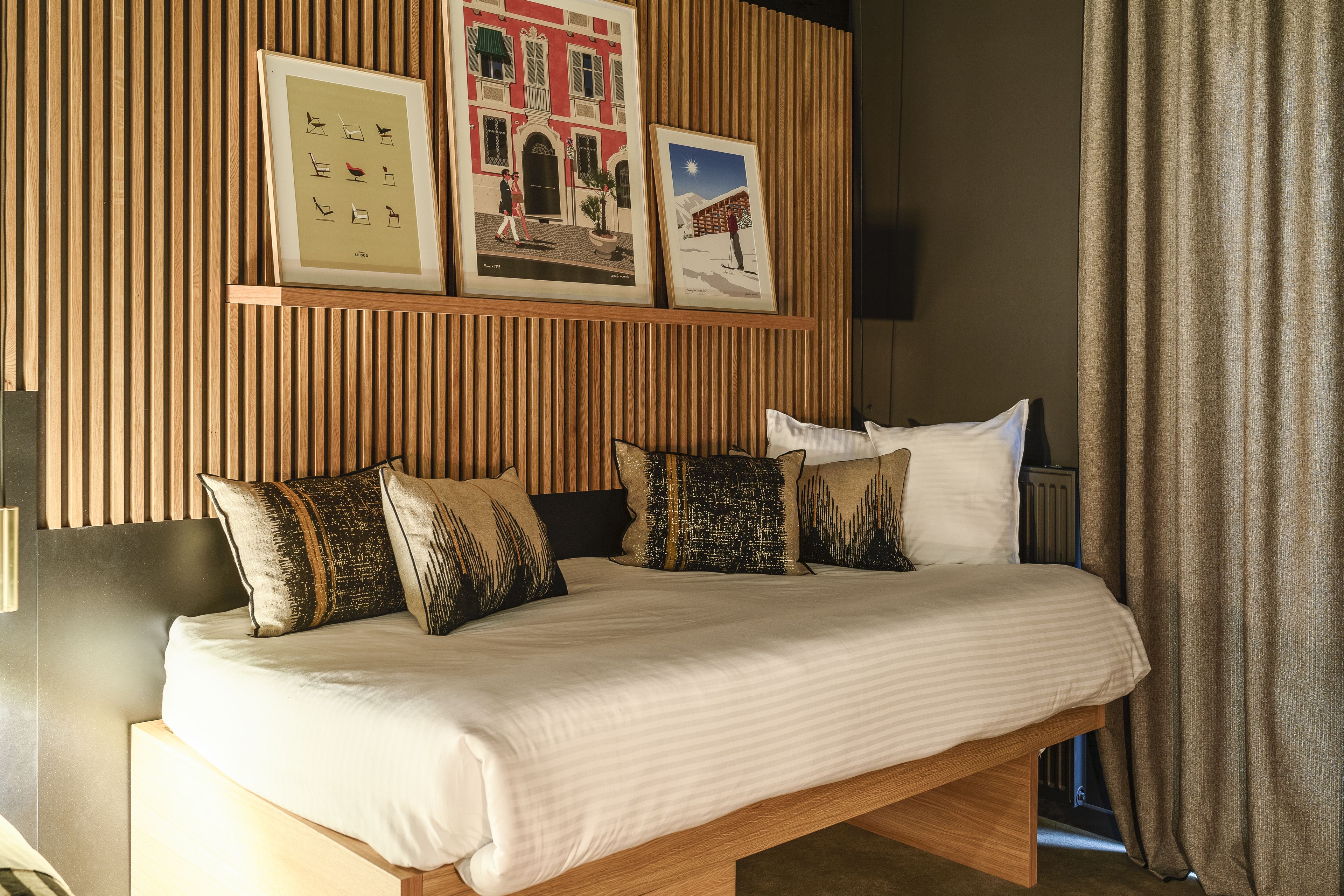 Una cama bien tendida con cubrecama blanco y a rayas, cabecera de madera y varios almohadones. En la pared sobre la cama hay obras de arte enmarcadas.